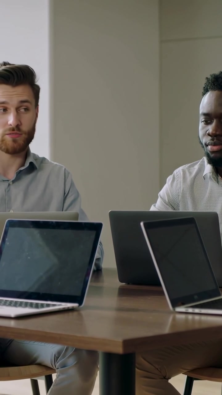 Vertical video: Glancing right, left man in shirt speaking during meeting, three laptops