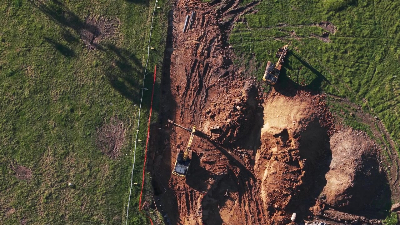 Drone video captures a bird's eye view of heavy excavators moving soil on a construction site surrounded by green grass in South East Melbourne, in striking afternoon light.