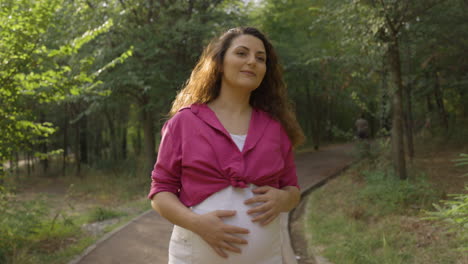 happy pregnant woman expectant mother walking alone in park turning around, attractive young casual dressed mom stroking caressing touching her belly with hands smiling, medium portrait shot