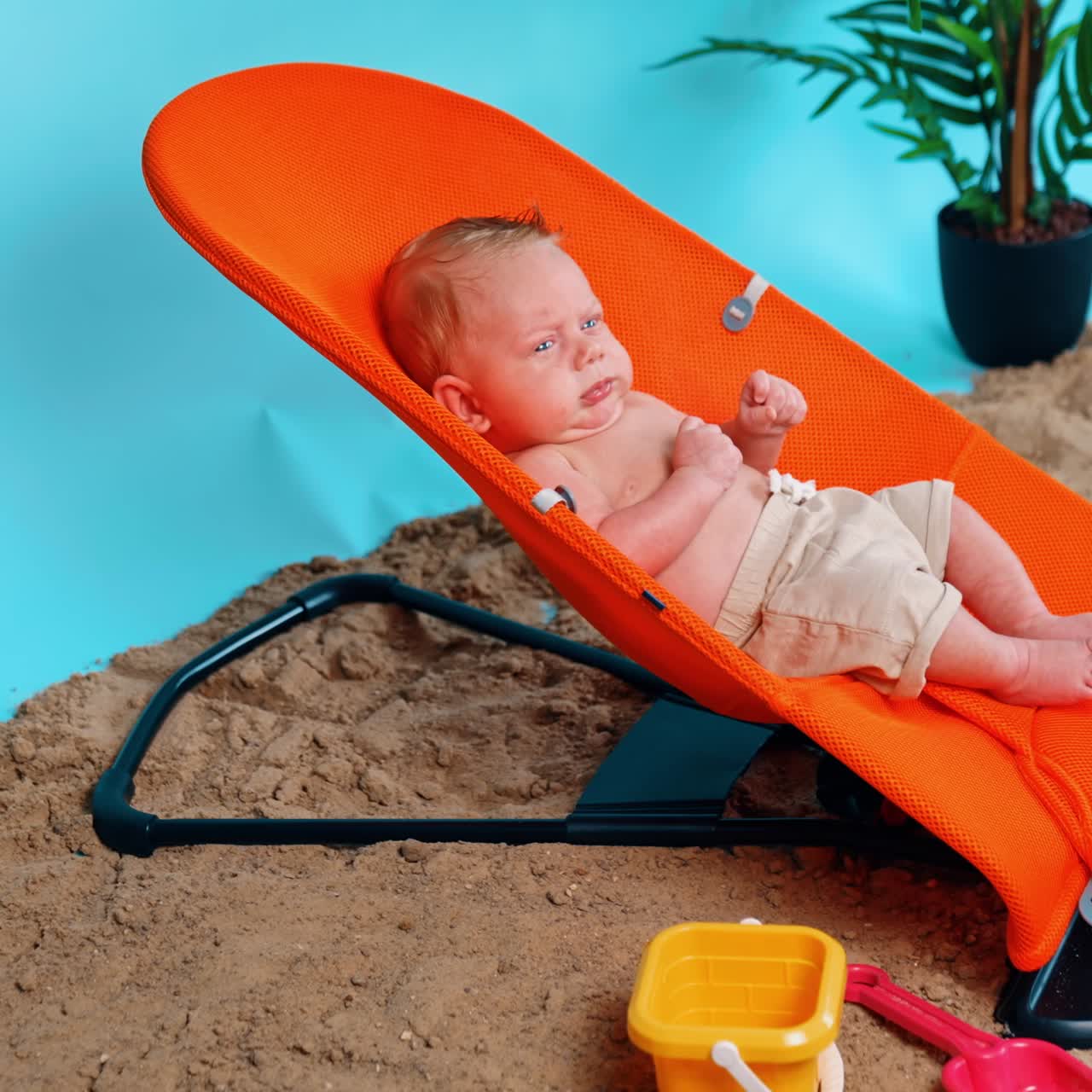 Nouveau-né sérieux en short assis dans une chaise pour bébé orange. Enfant se reposant sur une plage improvisée en studio