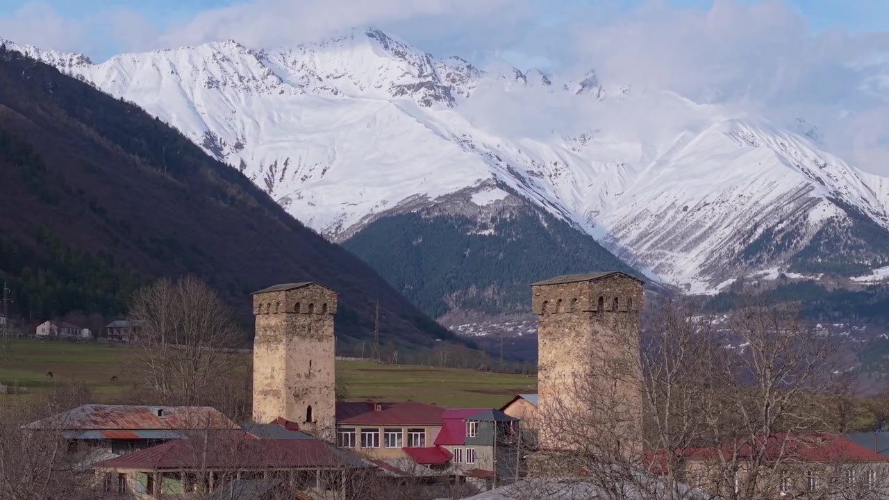 Órbita de drone com teleobjetiva em torno de duas torres Svan históricas em Mestia, revelando as majestosas Montanhas do Cáucaso cobertas de neve e a deslumbrante paisagem alpina da Geórgia