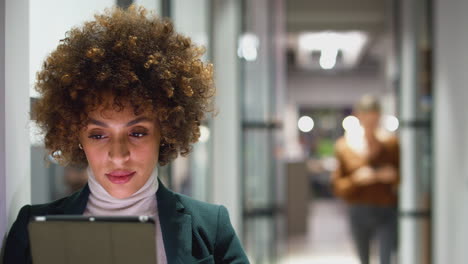 evening shot of businesswoman standing in corridor of modern office using digital tablet