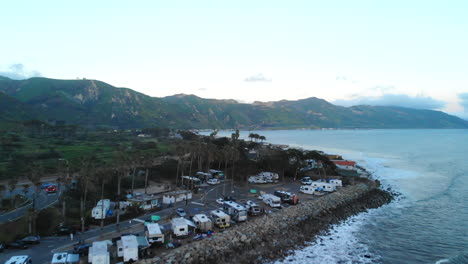 Aerial-drone-rising-over-an-rv-campground-on-the-beach-cliffs-of-Ventura,-California-with-Pacific-Ocean-waves-crashing-on-the-rocky-shore-below-at-sunset