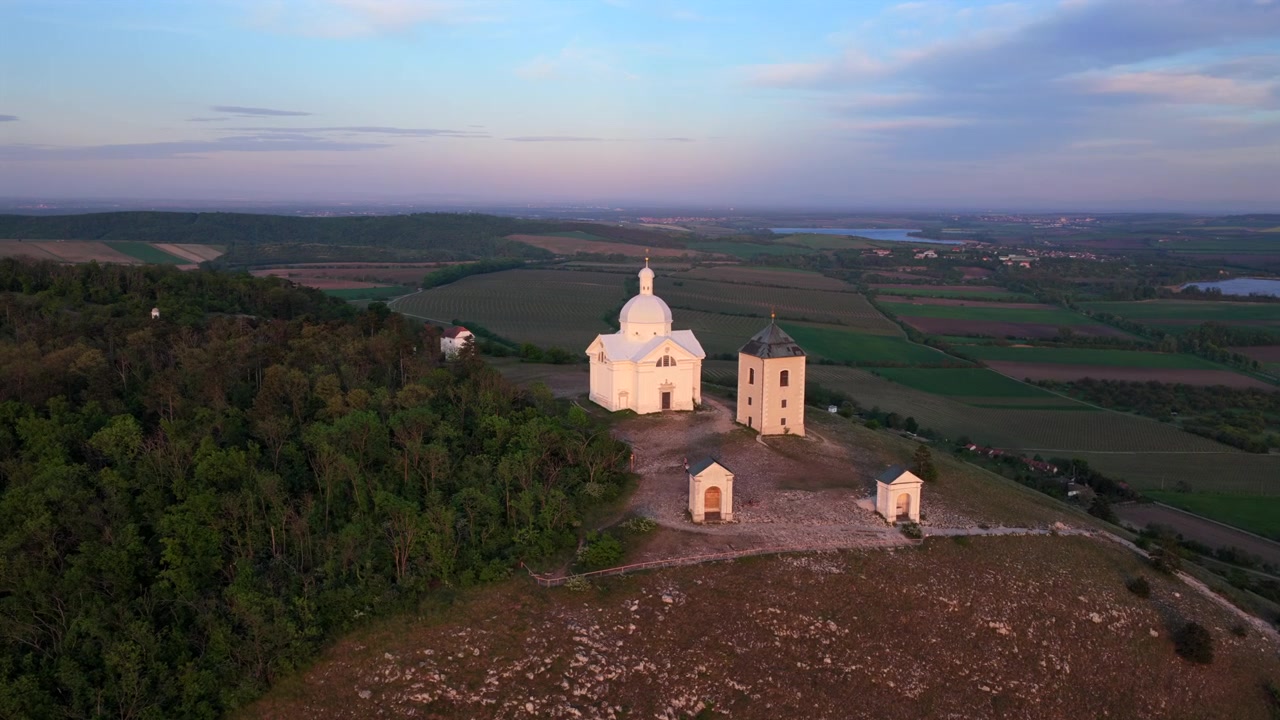Veduta aerea ad alta quota della Collina Sacra (Svatý Kopeček) con le sue cappelle della Via Crucis e la cappella barocca bianca sopra foreste e pianure di vigneti al tramonto, Mikulov, Moravia Meridionale, Repubblica Ceca.