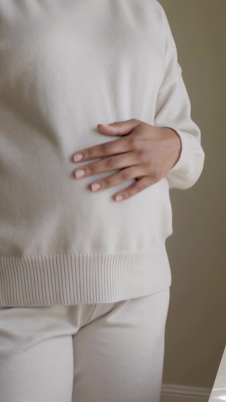 Vertical video: Lifting hand from glass, woman in cream sweater rubbing stomach for relief at home