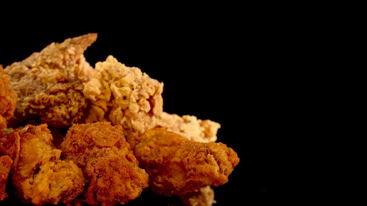 Crispy fried chicken breasts, thighs, and nuggets rotate on a black background under studio lighting, highlighting golden, spicy breading and texture