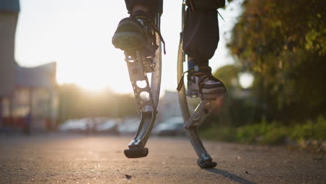 close-up of a person's leg walking on spring stilts with dark pants and sneakers. background shows a blurred suburban area with greenery, buildings, and vehicles under a warm sunset glow. low-angle