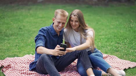 a guy and a girl on a picnic take a selfie. people sit on a mat in the park. happiness, leisure