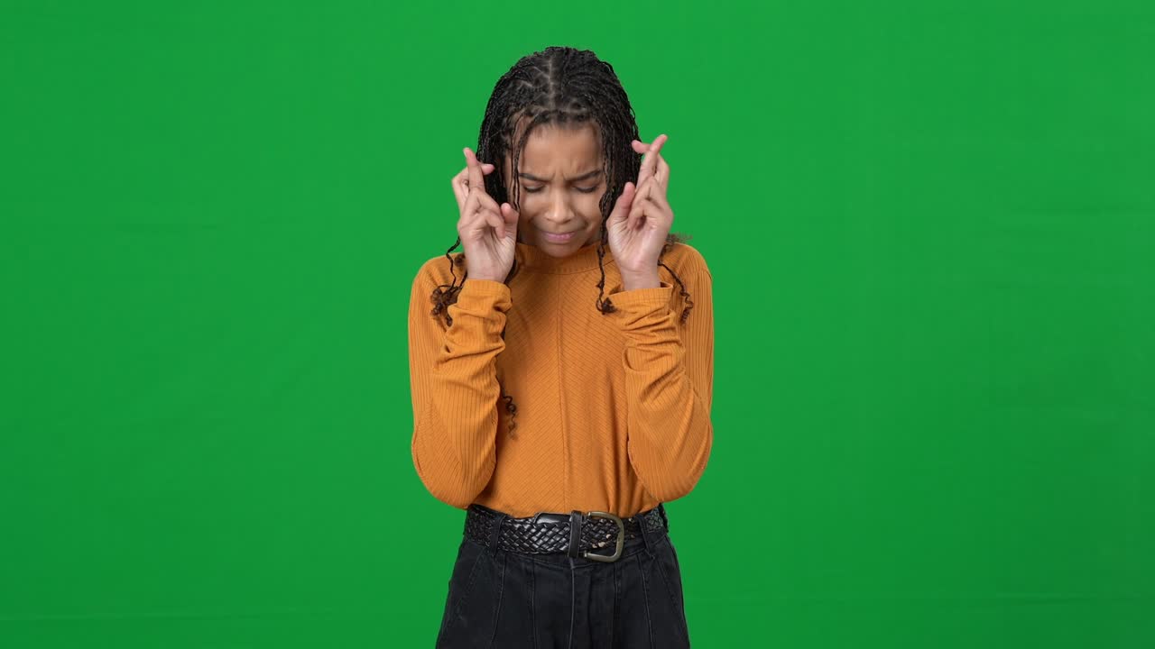 Excited teen African American girl crossing fingers closing eyes standing at chromakey background. Portrait of hopeful ambitious teenager waiting for test results or news on green screen. Hope concept.
