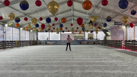 Niña-Patinando-Sobre-Hielo-Sola-En-Una-Pista-De-Hielo-Interior-Vacía-Con-Adornos-Navideños-Moviéndose-Hacia-La-Cámara