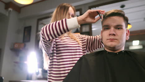 interior shot of working process in modern barbershop. side view portrait of attractive young man getting trendy haircut. male hairdresser serving client, making haircut using metal scissors and comb.