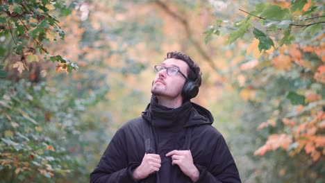 Young-European-man-with-headphones-in-a-vibrant-fall-mixed-forest,-observing-colorful-autumn-leaves