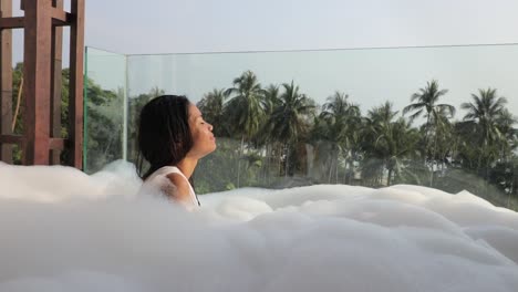 an asian woman taking a bubble bath in a jacuzzi on a balcony and taking a deep breath relaxing with a tropical view in the background