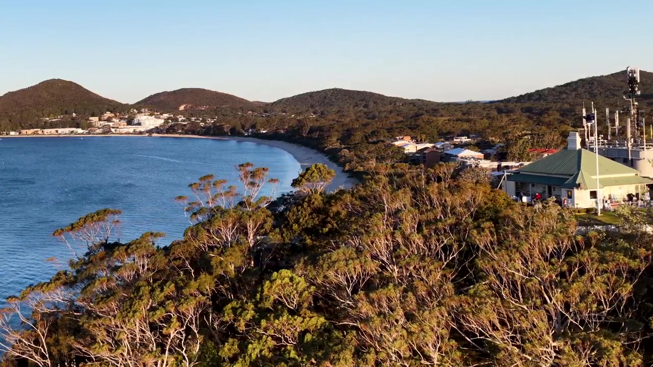 Tomada de drone em ângulo elevado fazendo panorâmica pela praia de Shoal Bay e colinas arborizadas em Nelson Bay, Austrália