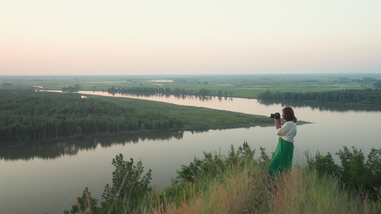 female photographer capturing distant river and forest landscape during sunset from cliff edge wearing green dress and white shirt, enjoying nature view with camera in hand against pastel sky