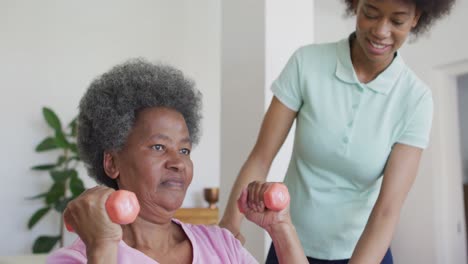 Happy-african-american-female-physiotherapist-helping-senior-female-patient-exercise-at-home