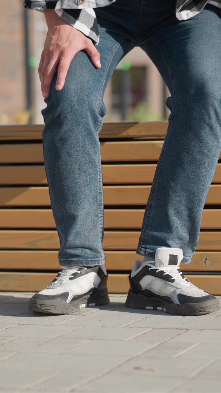 Primo piano di un uomo che indossa jeans blu e scarpe da ginnastica bianche, che cammina su un marciapiede vicino a panchine di legno. L'uomo si ferma e si siede, con le gambe visibili e le mani appoggiate sulle ginocchia, in un ambiente esterno con vegetazione.