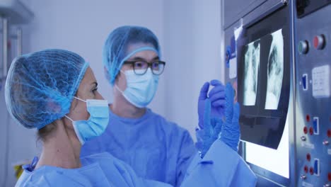 Diverse-male-and-female-surgeons-wearing-surgical-caps-and-face-masks-discussing-x-ray