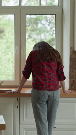 crying woman stands by kitchen window. brokenhearted wife hides tears leaning on countertop at home. lady deals with hard grief of husband death