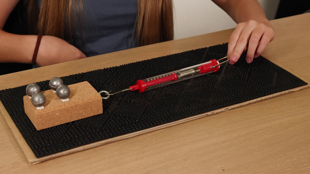 Hands use a spring scale to pull a weighted block across a textured surface, illustrating friction and force dynamics