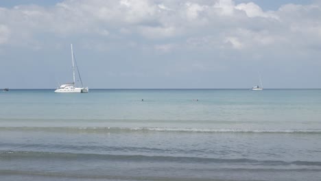 Der-Strand-Mit-Ein-Paar-Yachten-Im-Meer,-Weißer-Sand-Und-Wellen-Vom-Ruhigen-Meer-Bei-Sonnenschein-Tagsüber-In-Phuket