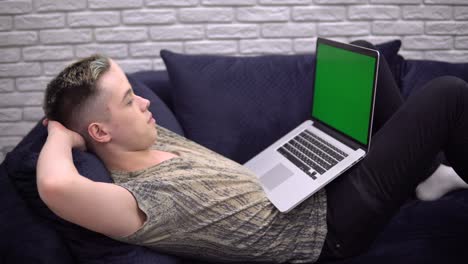 young student man relaxing on sofa, looking laptop green screen mockup