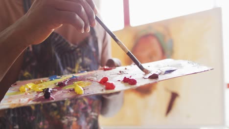 Mid-section-of-male-artist-wearing-apron-mixing-colors-in-a-palette-with-a-brush-at-art-studio