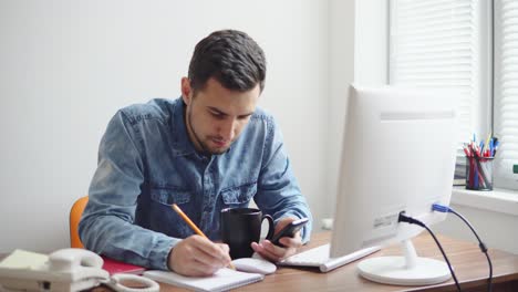 Young-businessman-sitting-by-the-computer-in-stylish-modern-office-and-taking-notes-using-his-pencil-and-phone.-Computer,-phone