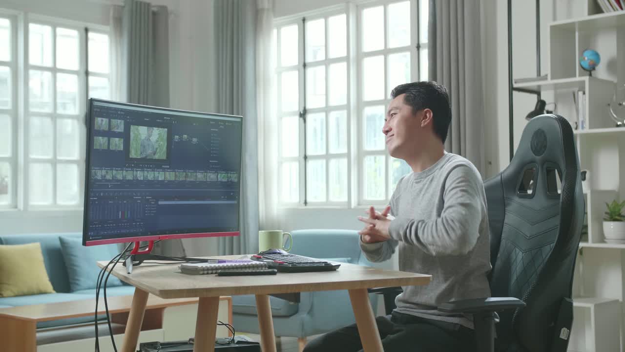 Asian Video Editor Man In Long Sleeved T-Shirt And Black Pants Stretching And Leaning Back While Using Desktop Computer For Editing Photo At Home.
