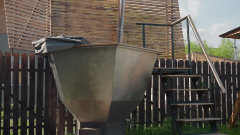 backyard view featuring large metal tub with vertical chimney and staircase beside wooden swing and tall dark fence, gentle sunlight casting soft shadows and green foliage