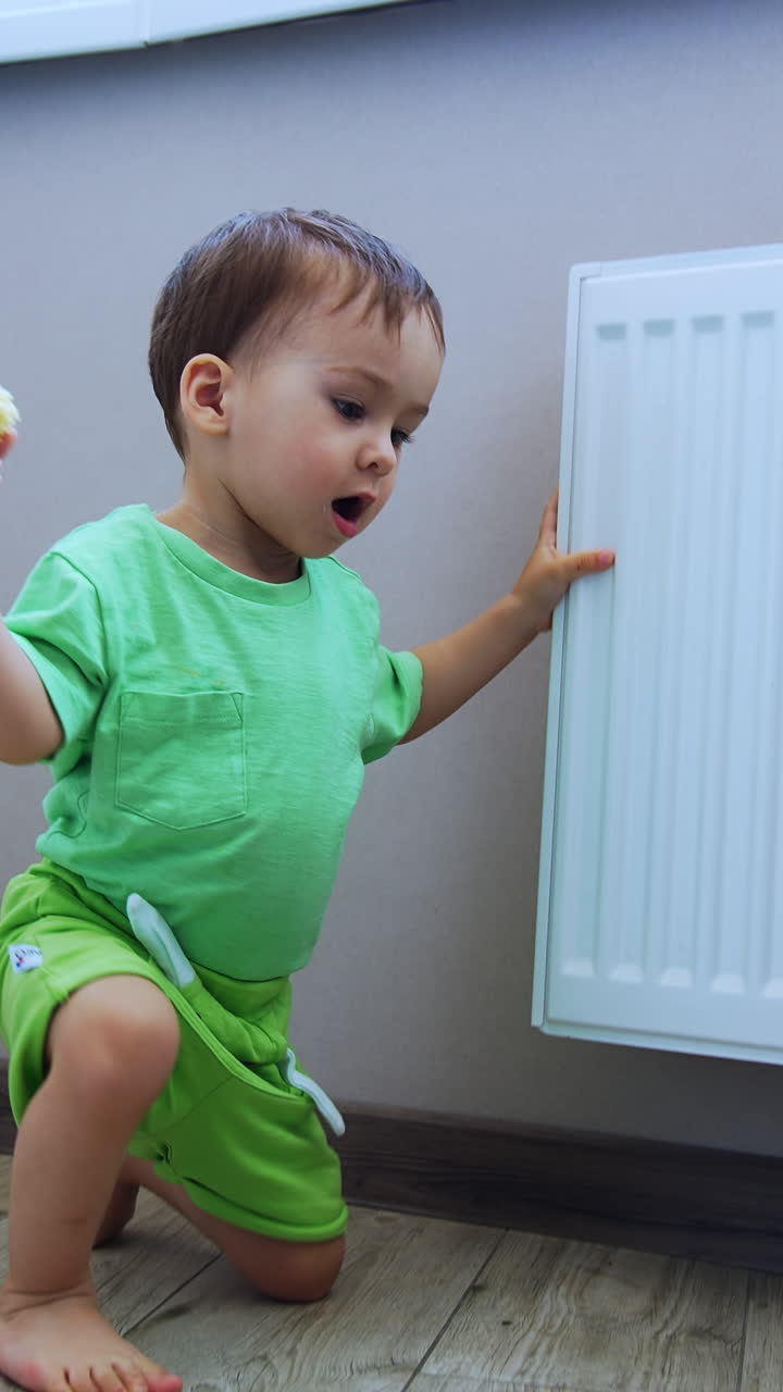 Toddler in green t-shirt and shorts sits on the floor near the radiator. Baby with food in hand stands up and walks by the room. Low angle view. Vertical video