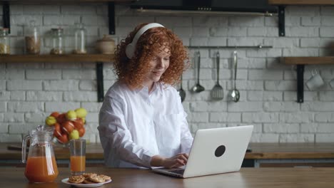 mujer roja sonriente joven con cabello rizado poniendo auriculares mientras trabaja en una computadora portátil en una cocina acogedora en casa. trabajo a distancia desde casa