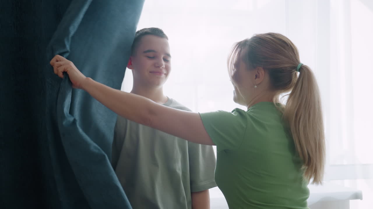Guardian discovers younger hiding behind curtain during hide and seek, touches nose with gentle gesture, playful moment transforms into hug, sunlight through window