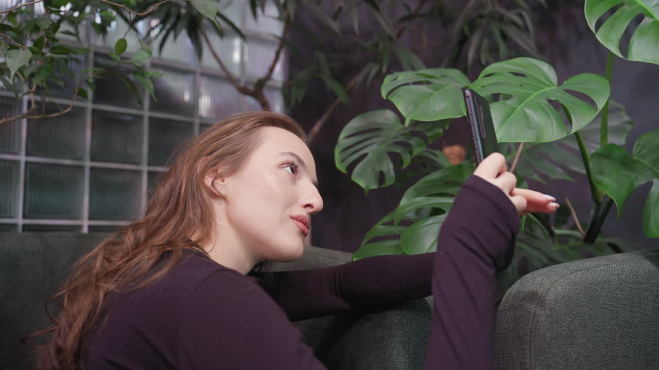 Blond woman with soft smile leans on green cushion chair while concentrating on taking photo with phone, surrounded by lush tropical plants and cozy indoor natural light setting