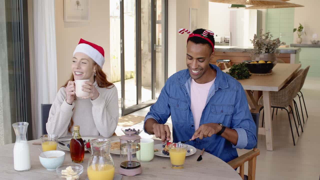 Diverse couple enjoying festive breakfast at home, wearing Christmas hats, smiling
