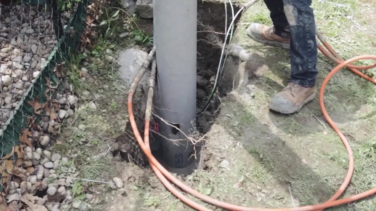 Un operaio utilizza un compattatore idraulico per compattare il terreno attorno alla base di un palo di illuminazione stradale appena installato, mentre cavi arancioni, terra esposta, rete protettiva e uno strumento di scavo incorniciano la fondazione attiva