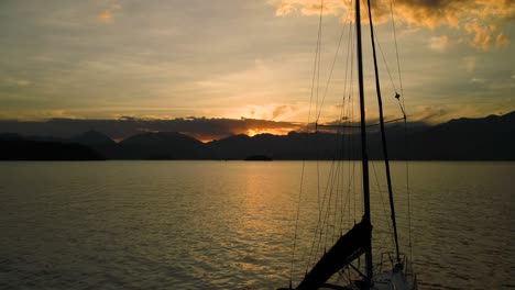 beautiful aerial drone subtle traveling up of sail boat in calm ocean with amazing orange sunset with clouds and mountains showing the mast and then only the background