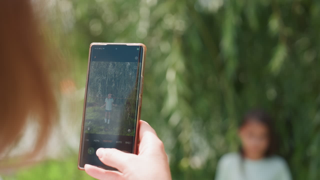 Mamma caucasica fotografa la figlia latina accanto a un salice; sessione di ritratto all'aperto con smartphone in primo piano, mano sfocata, bokeh soffice, luce solare estiva, espressione serena, legame materno