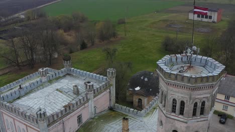 Tower-Of-An-Old-Neo-Gothic-Manor-Mansion-With-Latvian's-National-Flag-Waving-Against-Meadow-Countryside