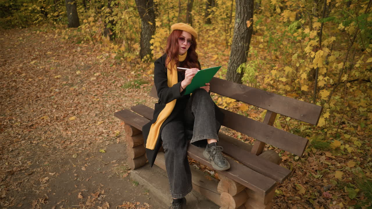 Vista dall'alto di un'artista che disegna pensierosamente mentre è seduta su una panchina di legno del parco in un tranquillo ambiente autunnale, con una gamba sulla panchina e una a terra, circondata da foglie dorate.