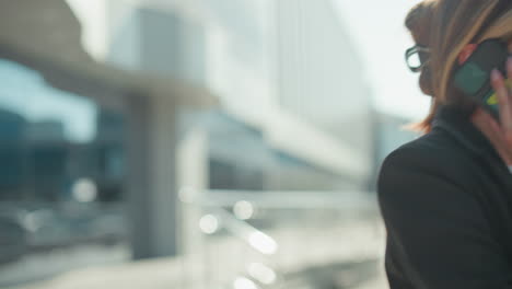 calm lady in professional outfit walks home after work while talking on phone, holding conversation with composed expression outside modern glass building reflecting cityscape under daylight
