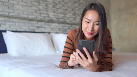 Mujer-Asiática-Sonriente-Acostada-En-La-Cama-Y-Pasando-El-Dedo-Por-La-Pantalla-Del-Teléfono-Móvil,-Con-La-Cara-Cerrada