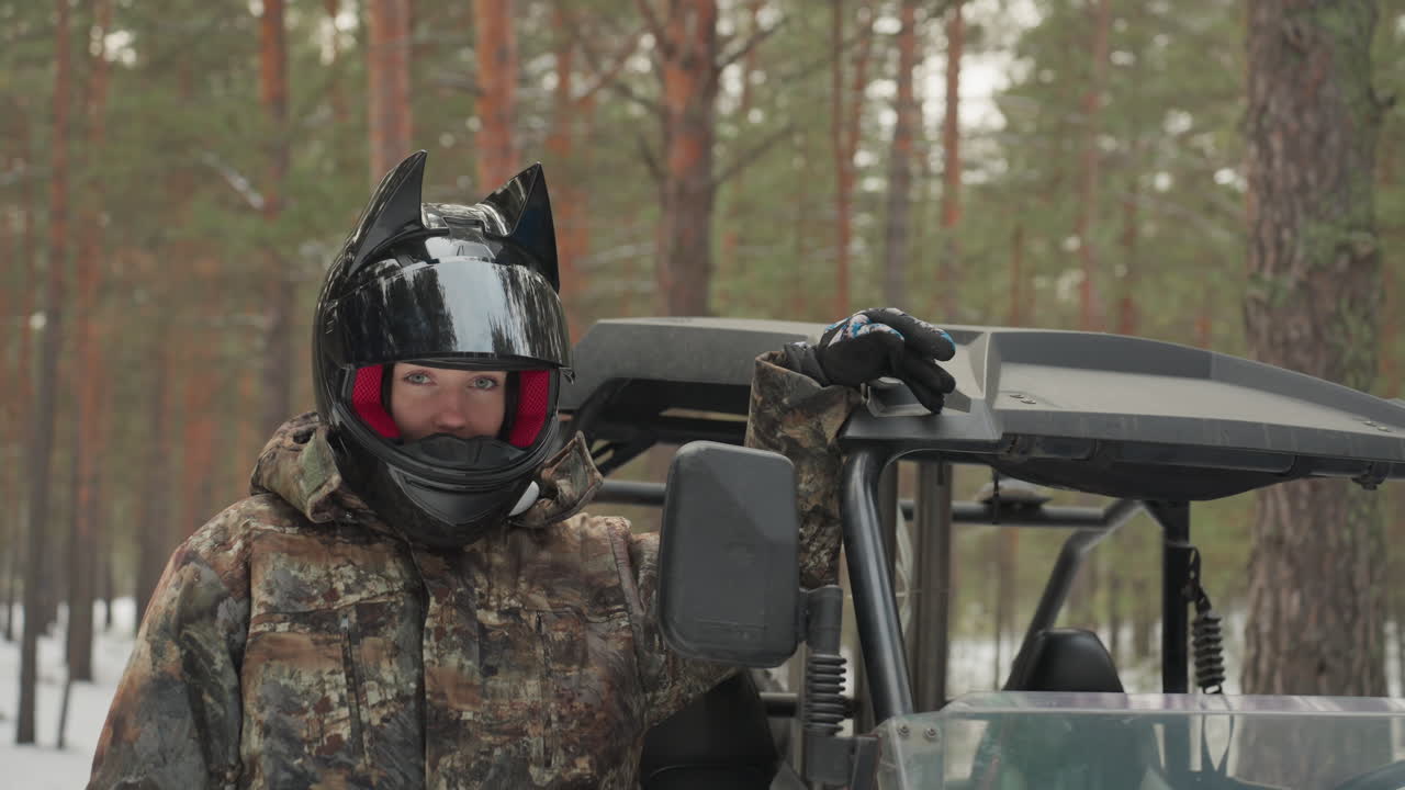 Pilota di ATV nella foresta innevata con casco a orecchie da gatto che stabilizza la portiera di un UTV, giacca mimetica e passamontagna rosso, leggera neve sui pini, pausa avventurosa e giocosa prima di una corsa fuoristrada, mano sullo specchietto e casco vicino.