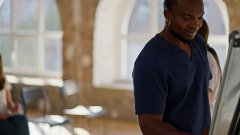 Close-up-shot:-A-Black-male-doctor-in-a-blue-medical-uniform-writes-in-felt-tip-pen-on-canvas-as-a-nurse-conducts-medical-training-in-the-background