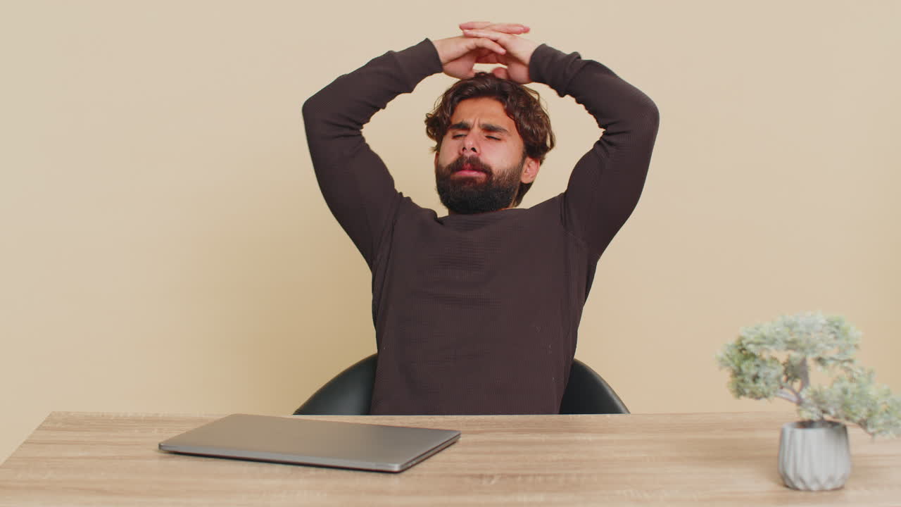 Bored indian young man freelancer closing laptop and sitting at table against beige background