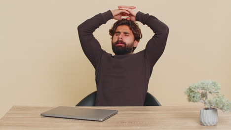 bored indian young man freelancer closing laptop and sitting at table against beige background