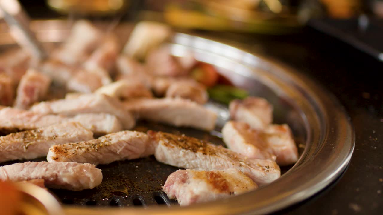 Close-up of pork grilling on metal pan, tongs turning meat, warm lighting, shallow focus