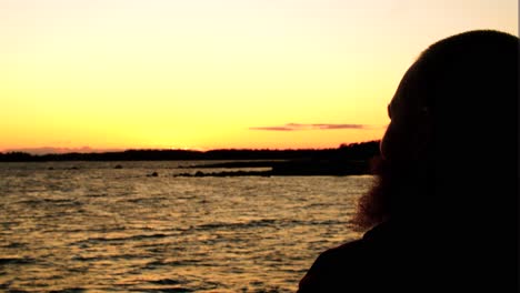 silueta de un hombre con barba fumando una pipa sosteniéndola fuertemente humo saliendo de la boca mirando alrededor sobre el horizonte de las aguas corriendo corrientes de olas delante de él puesta de sol