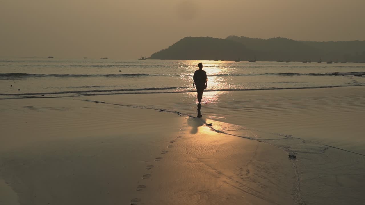 Landscape view of a young woman walking on a sandy beach by the ocean, at sunset