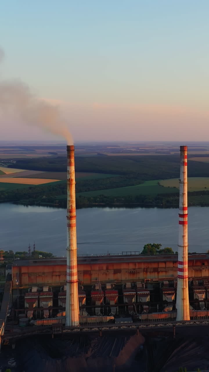 Chemical plant among beautiful nature. Industrial factory with smoking pipe near the river at sunset. Pipe emits harmful emissions and pollute the environment. Aerial view. Vertical video